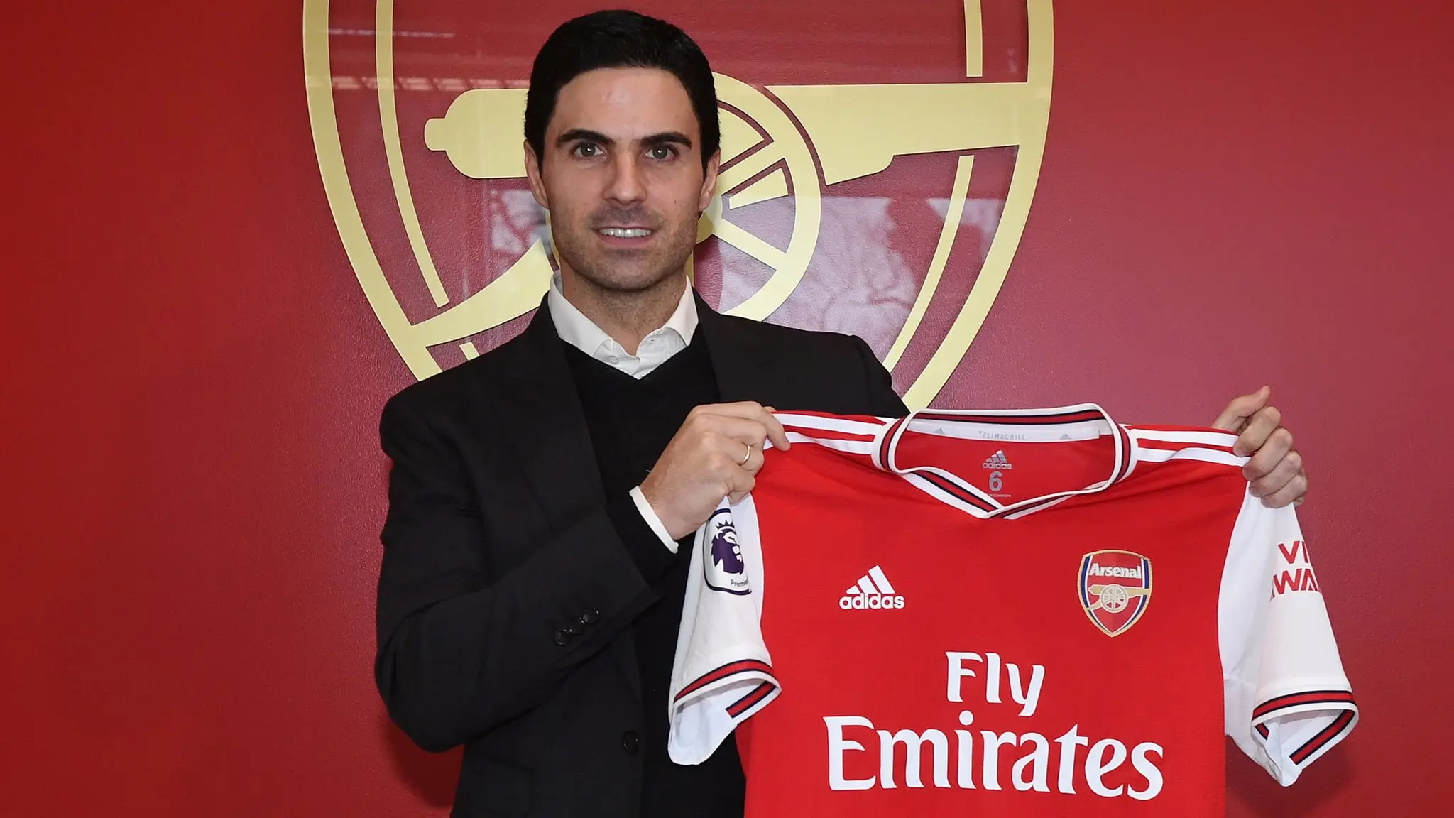 Mikel Arteta holding an Arsenal jersey in front of the club logo.