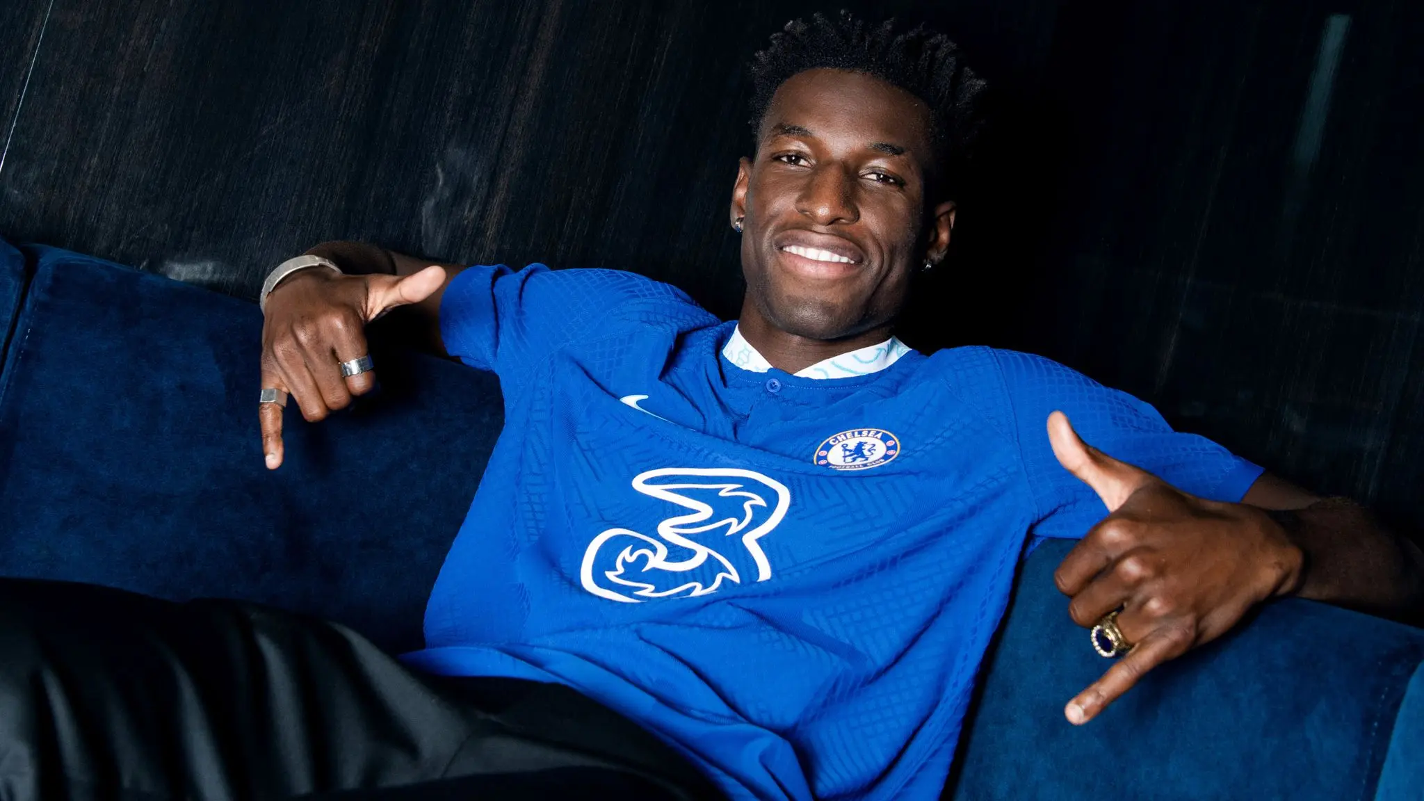 Nicolas Jackson posing in a blue Chelsea football jersey on a sofa.
