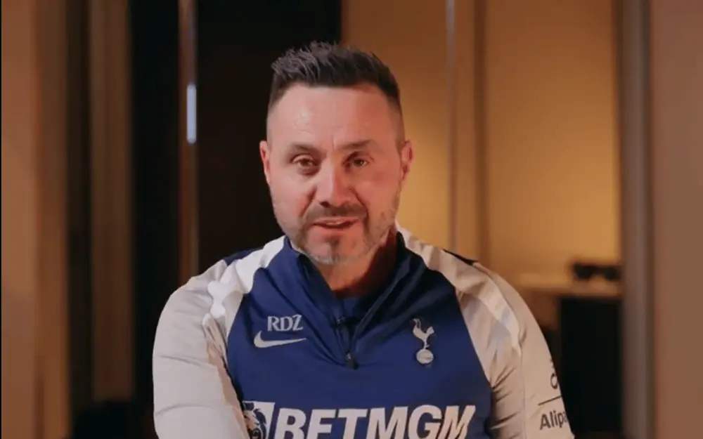 Roberto De Zerbi wearing a Tottenham Hotspur training shirt indoors.